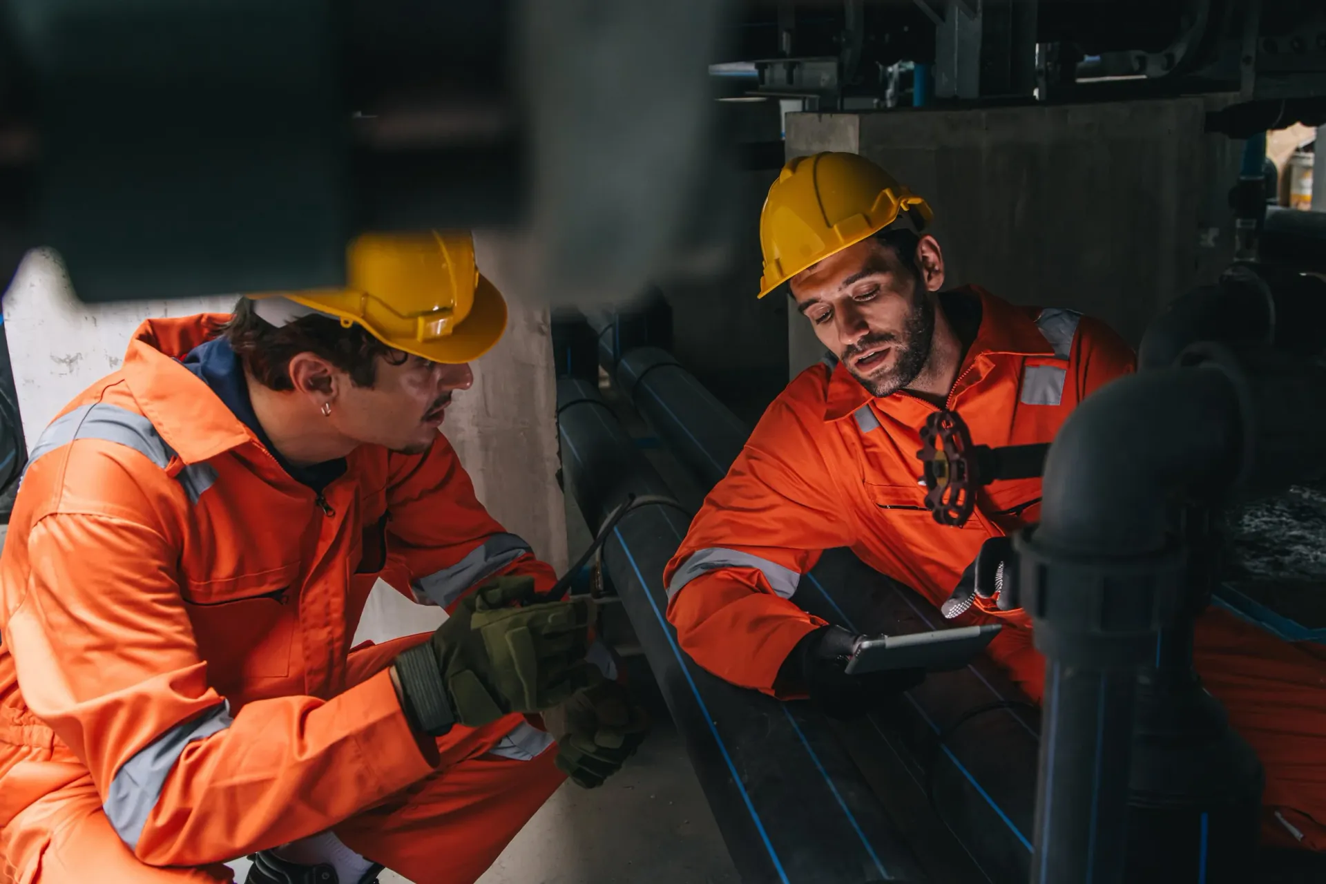 Two workers in orange high-visibility overalls and hard hats crouched over industrial pipework, one holding a tablet